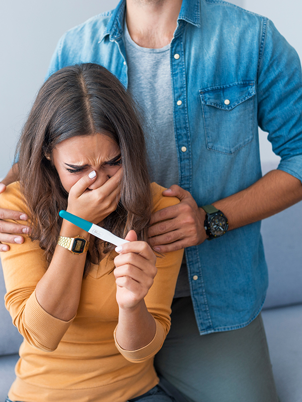 upset woman looking at a pregnancy test while behing comforted by a man behind her